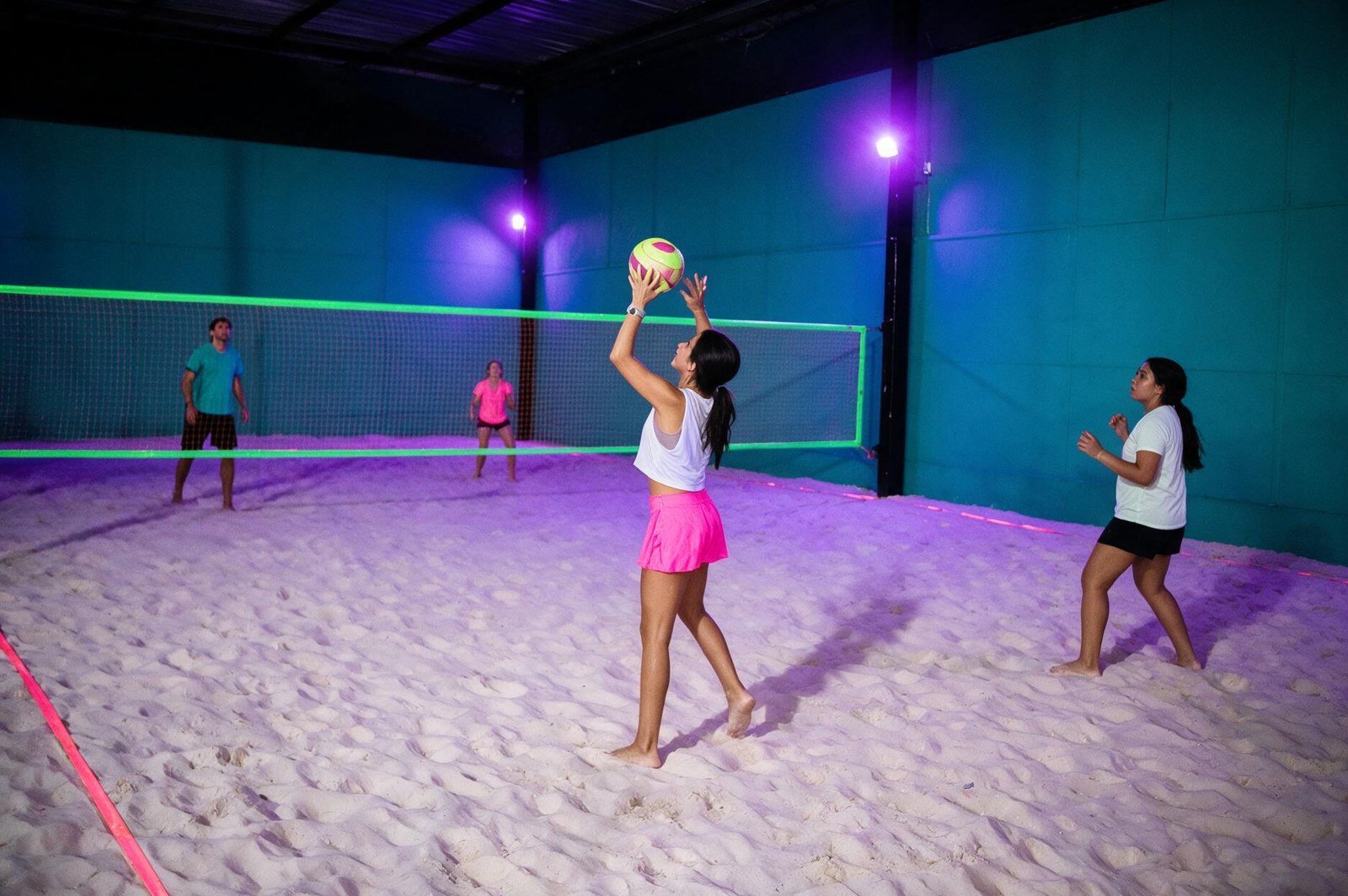 Partida noturna na quadra de areia da Tropical Beach sob iluminação neon roxa, com rede em fita verde fluorescente