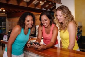 Amigas sorrindo em pé no bar da Tropical Beach escolhendo no celular entre quadra avulsa e plano mensal